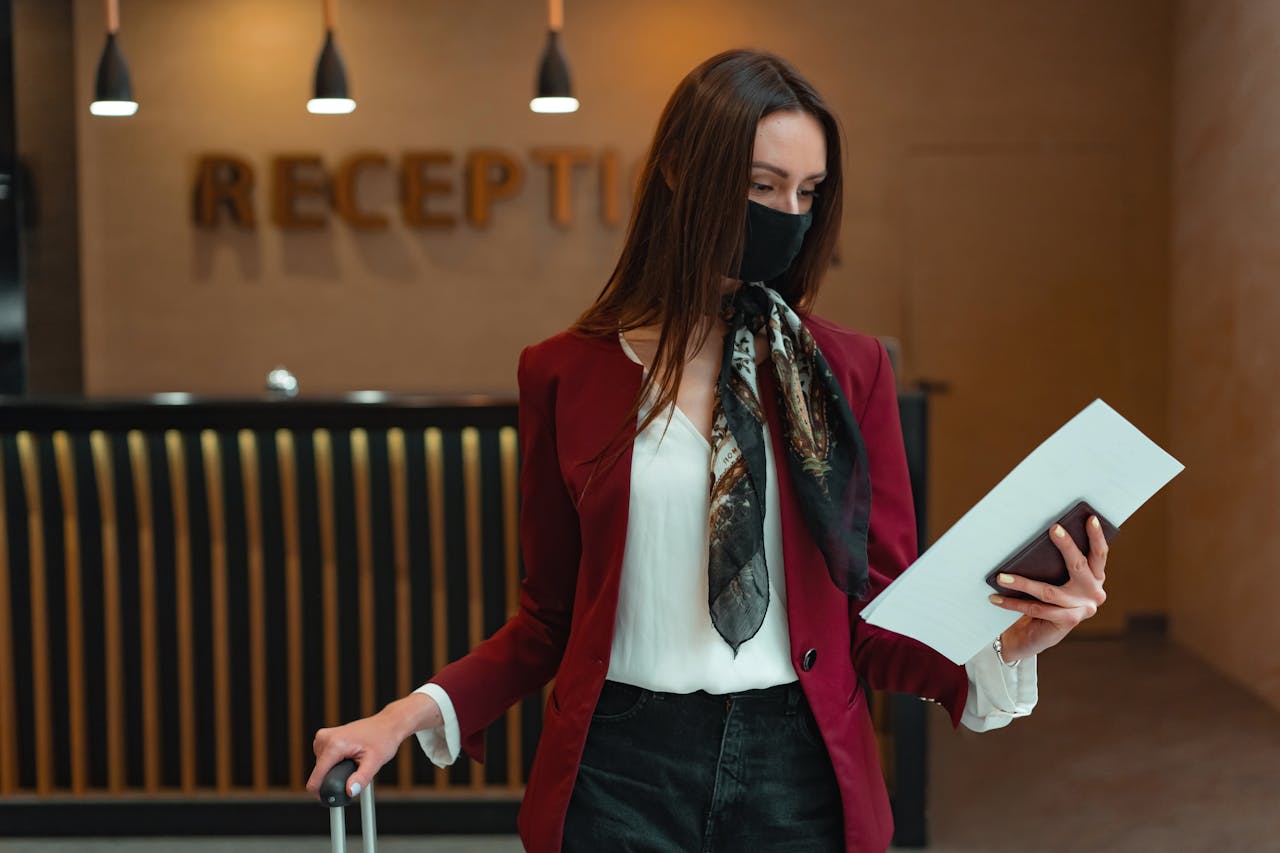 Woman in red blazer at hotel reception, holding luggage and documents, wearing a face mask.